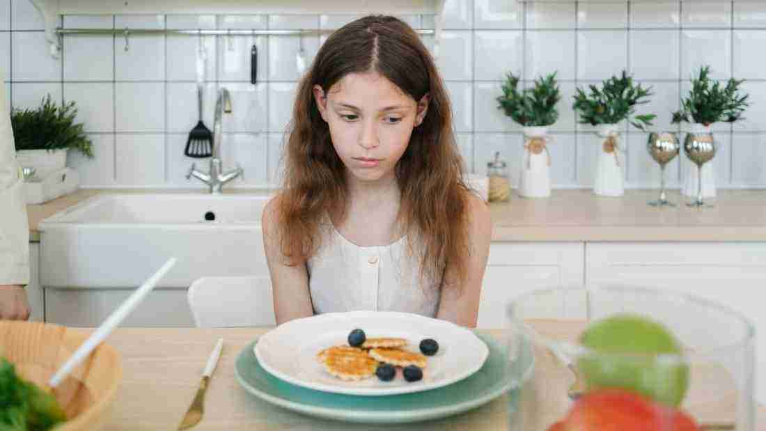 A sad little girl sitting in front of a plate of food that she is not eating