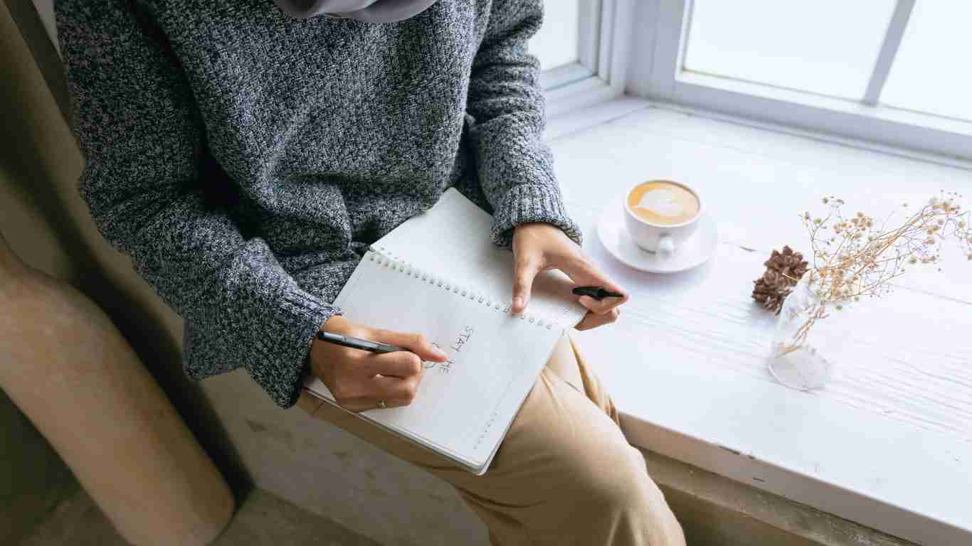 A woman sitting on a window sill and writing in her journal