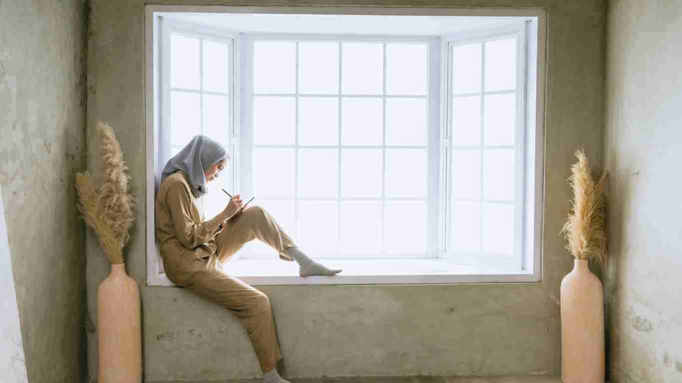 A young woman in a hijab sitting on a windowsill and writing in her journal